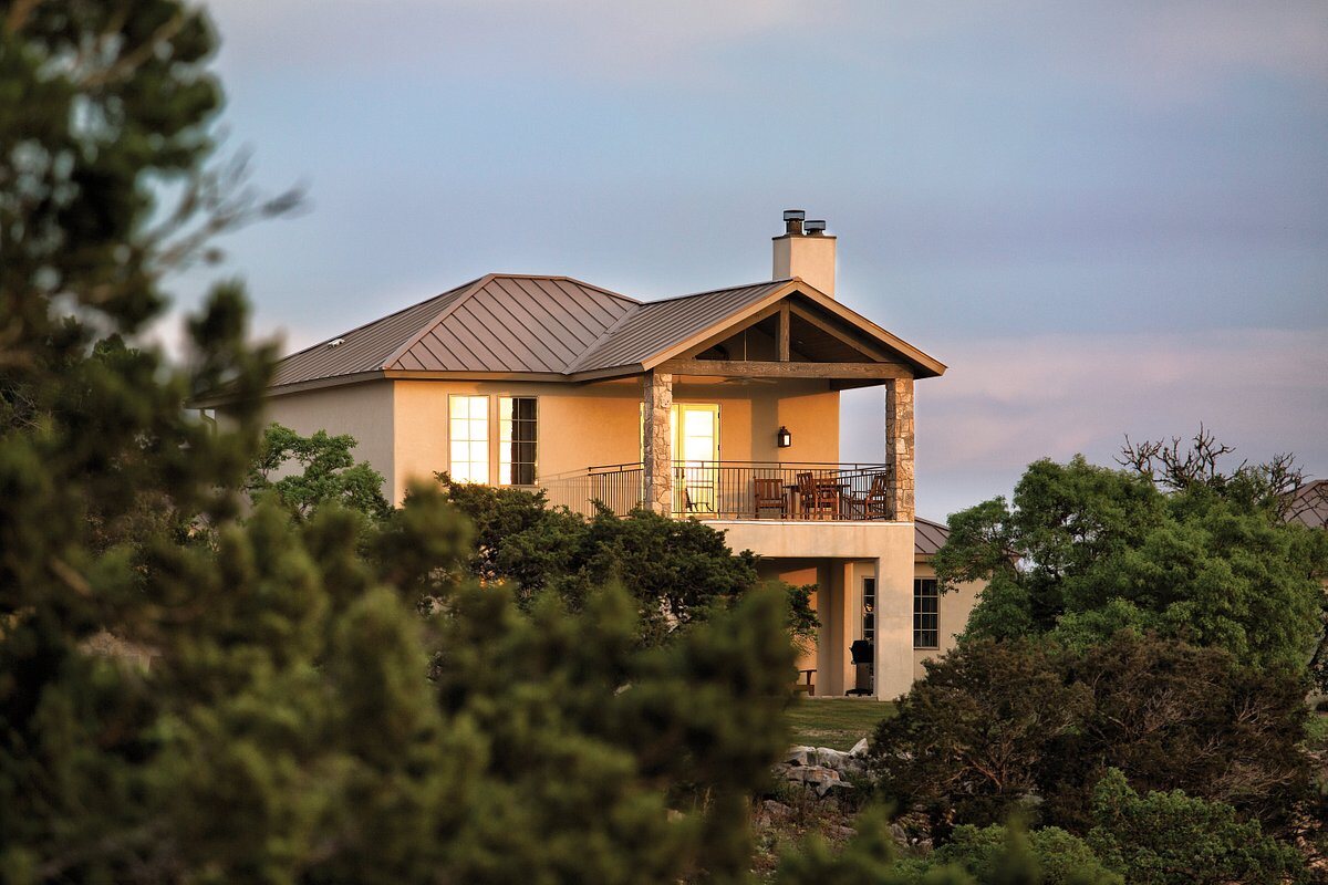 Two-story stucco CEB home with a metal roof and covered balcony on a Hill Country hillside